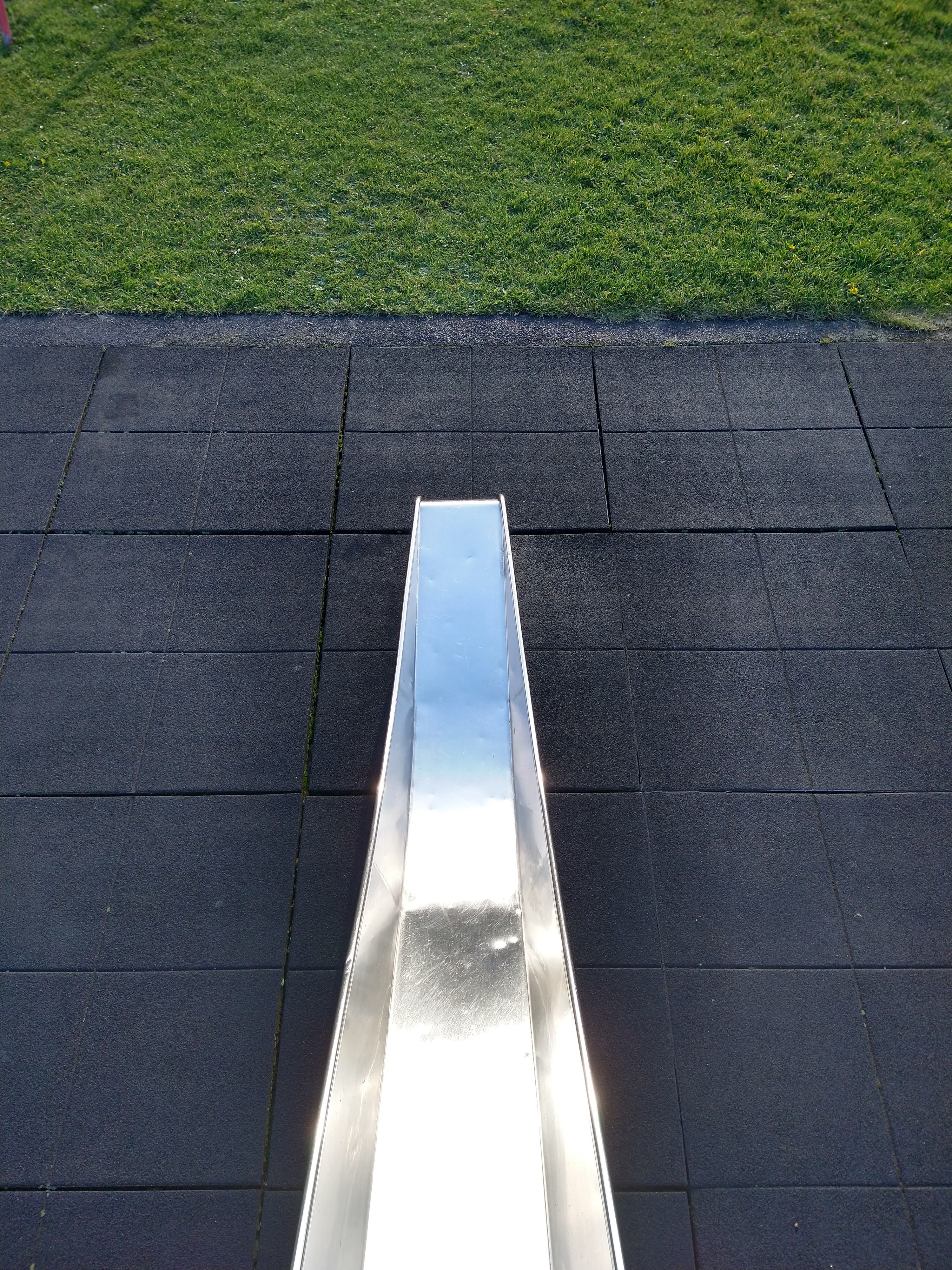 A picture of a silver slide in the middle on black rubber floor with green grass at the top of the screen